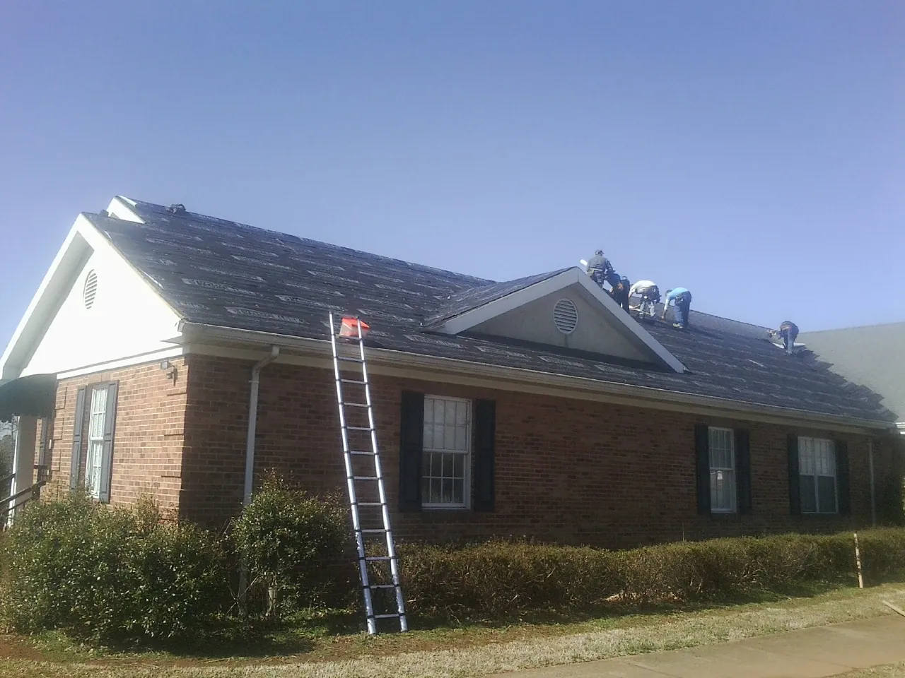 Crew tearing shingles off a brick ranch roof with a ladder set — Metal Roof Repair work by our crew in Holly