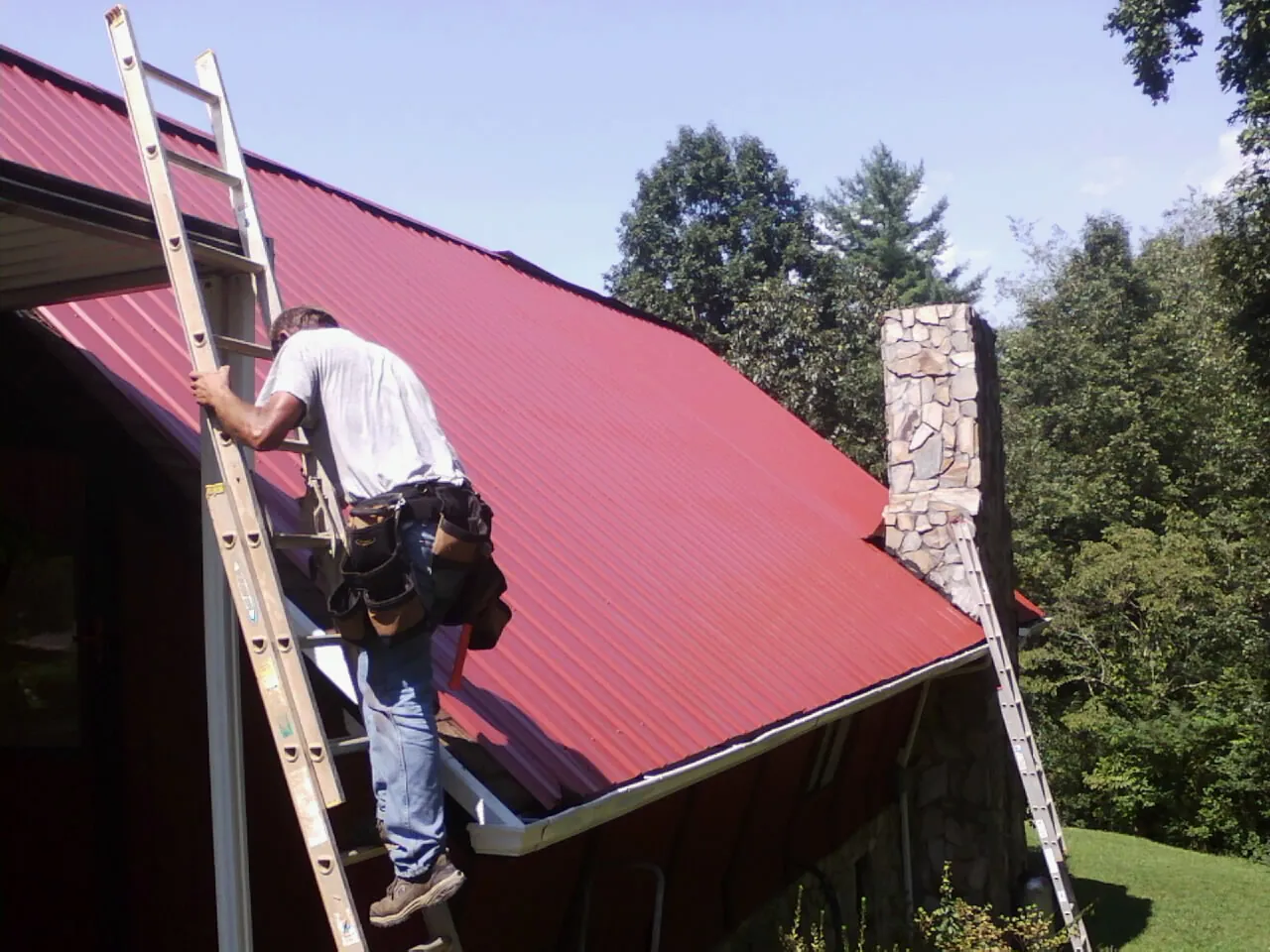 Roofer on a ladder beside a red metal roof and stone chimney — Soffit & Fascia Repair by Roofing Brothers in Holly